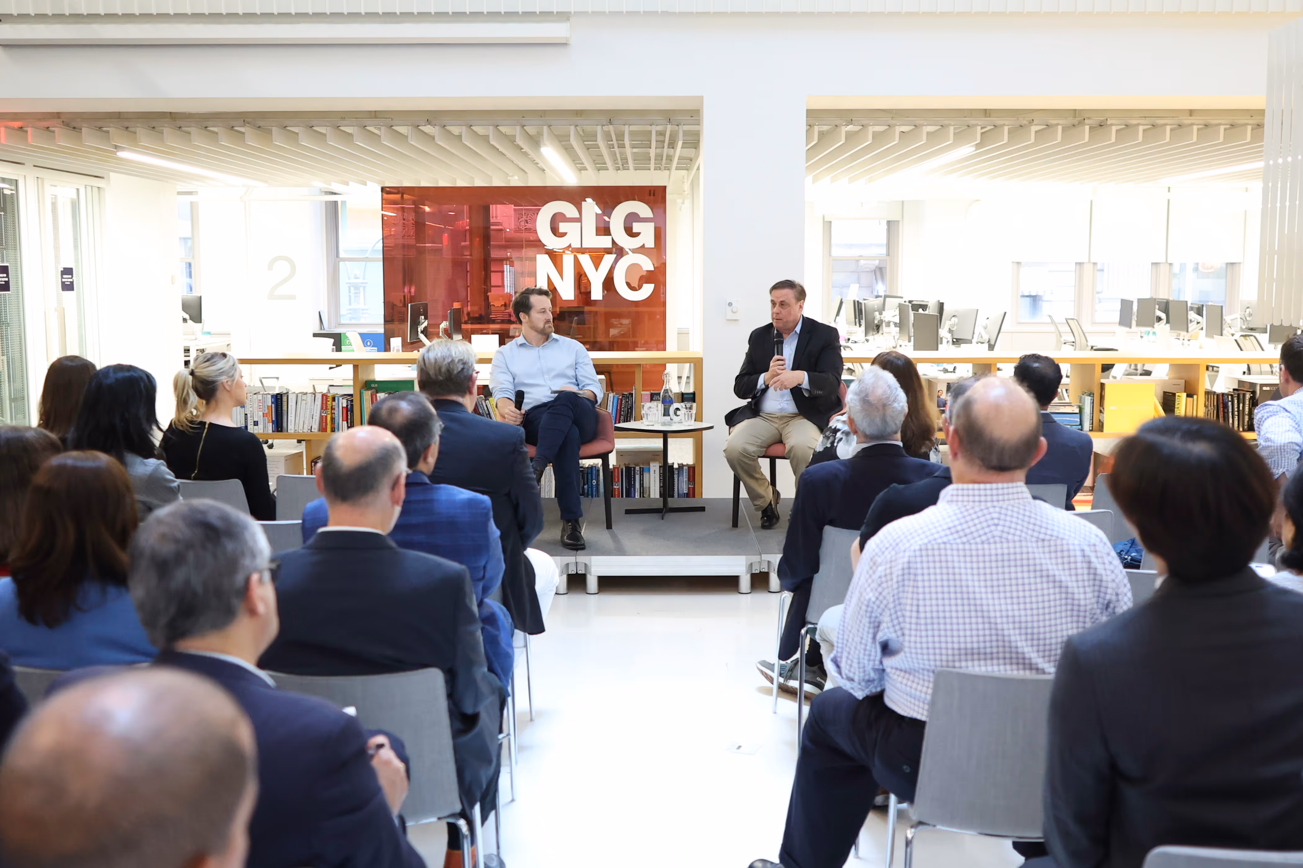 Two men seated on a small stage having a discussion with an audience in a bright modern office space with GLG NYC sign in the background.