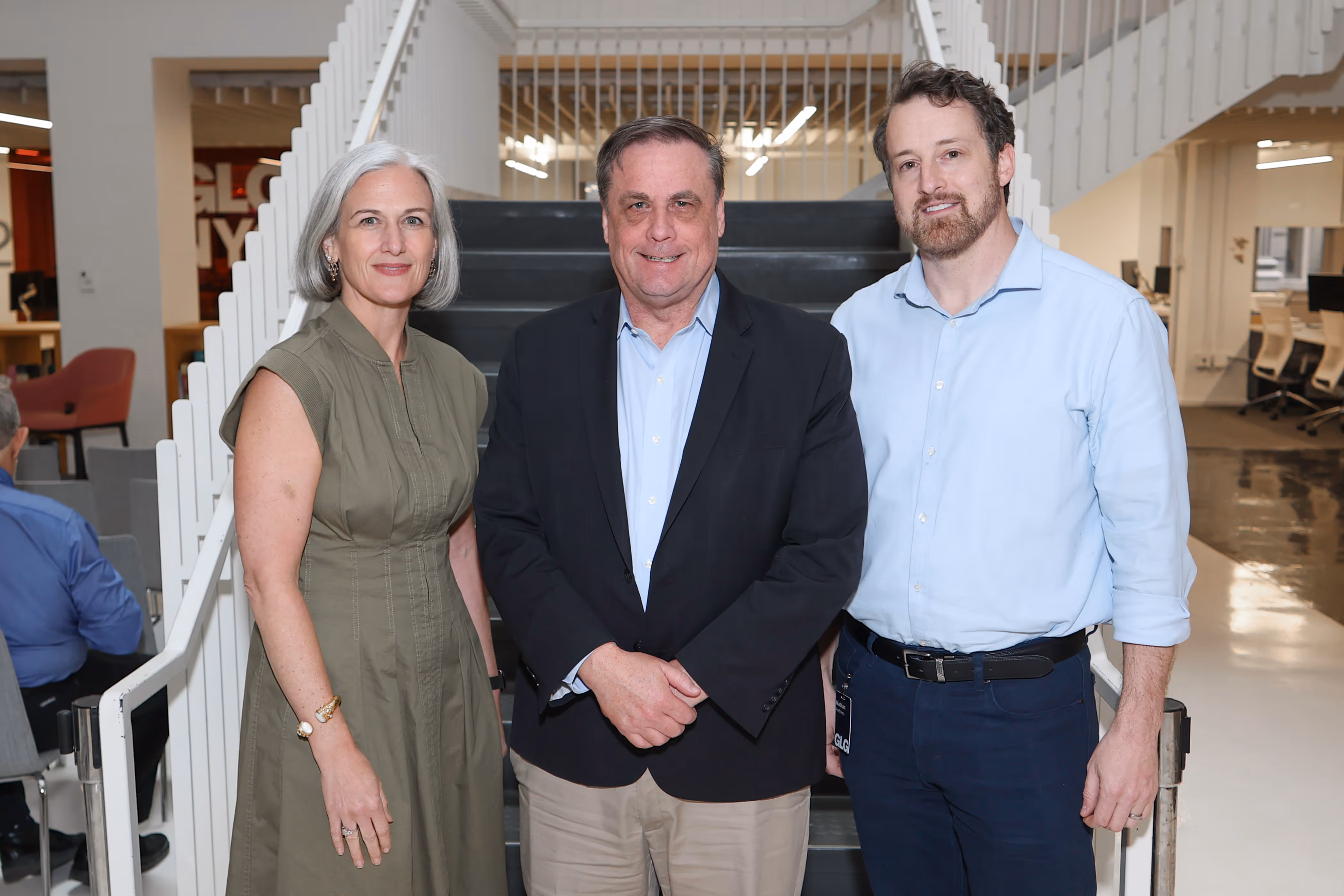 Three people standing indoors in front of black stairs, two men and one woman, all smiling at the camera.