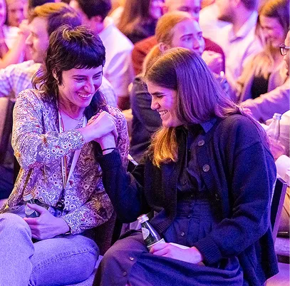 Two women smiling and fist bumping in a crowded, warmly lit social gathering.
