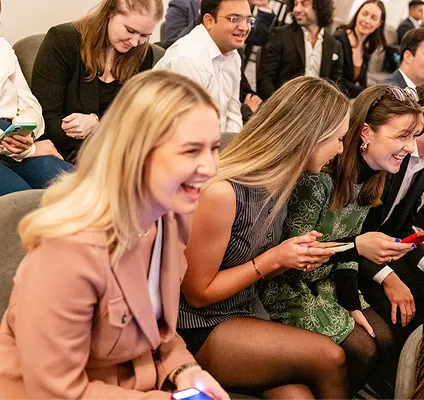 Group of young adults sitting closely, smiling and laughing while holding smartphones indoors.