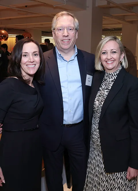 Three people smiling, two women in dresses and a man in a suit at an indoor event.