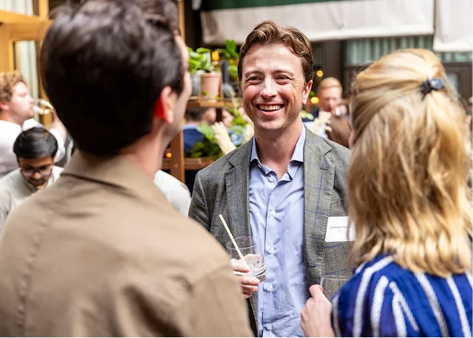 Smiling man holding drink and talking with two people at a social gathering indoors.