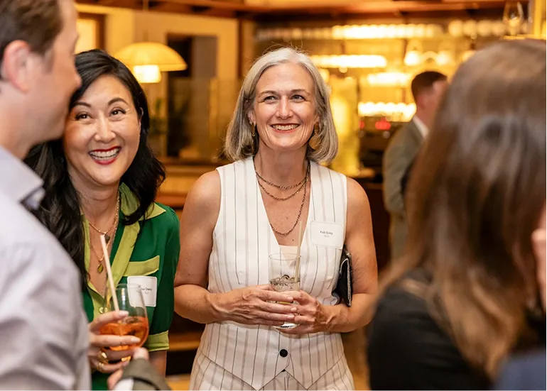 Two women smiling and holding drinks, socializing at an indoor event with blurred people background.