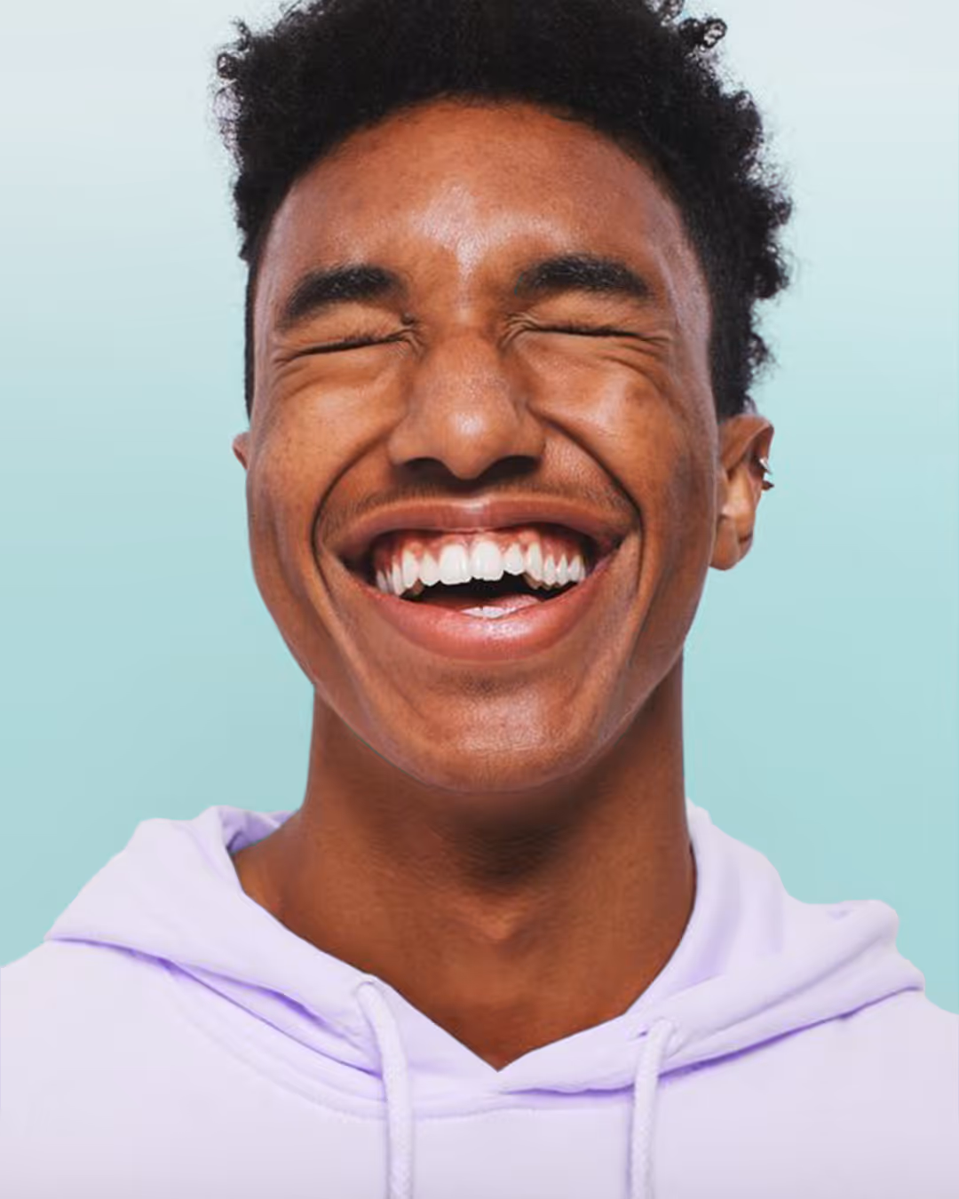 Jonge man met krullend haar en een lichtpaarse hoodie lacht met gesloten ogen en brede glimlach.