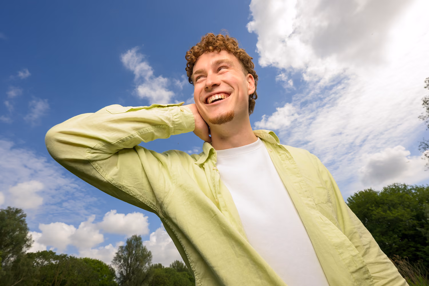 Jonge man buiten lacht en kijkt omhoog, draagt lichtgroene blouse en witte T-shirt onder blauwe lucht.