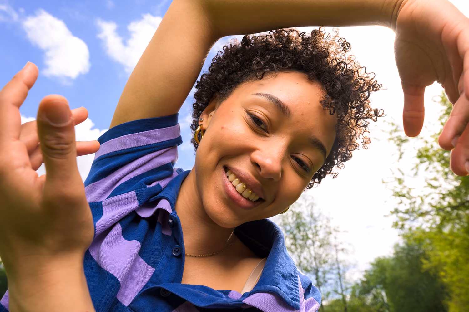Lachende vrouw met krullend haar in blauw-paarse gestreepte polo poseert buiten onder blauwe lucht.