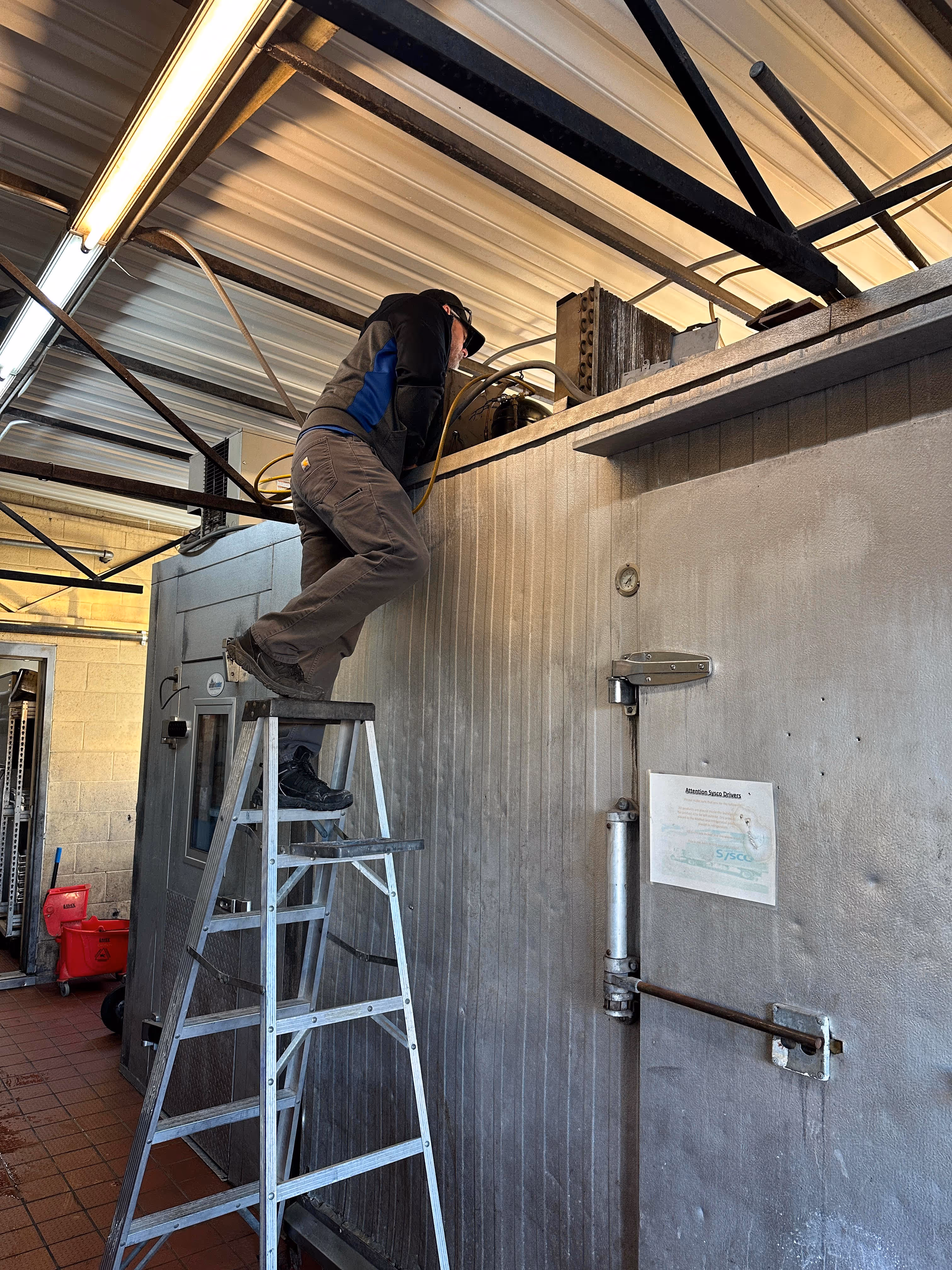 Person standing on an aluminum step ladder working on industrial refrigeration equipment inside a facility.
