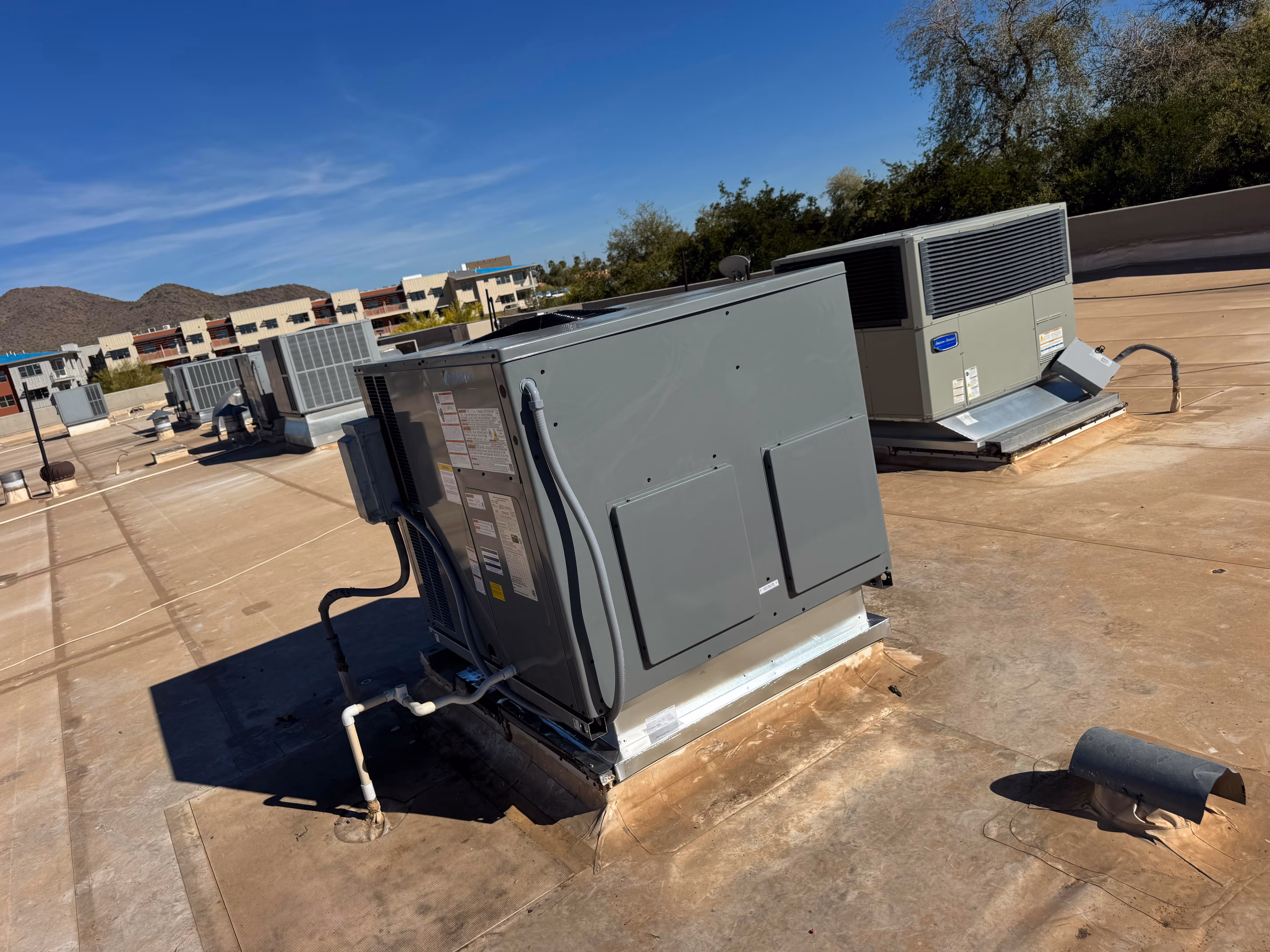 Multiple rooftop commercial HVAC units installed on a flat roof with distant mountains and buildings under a clear blue sky.