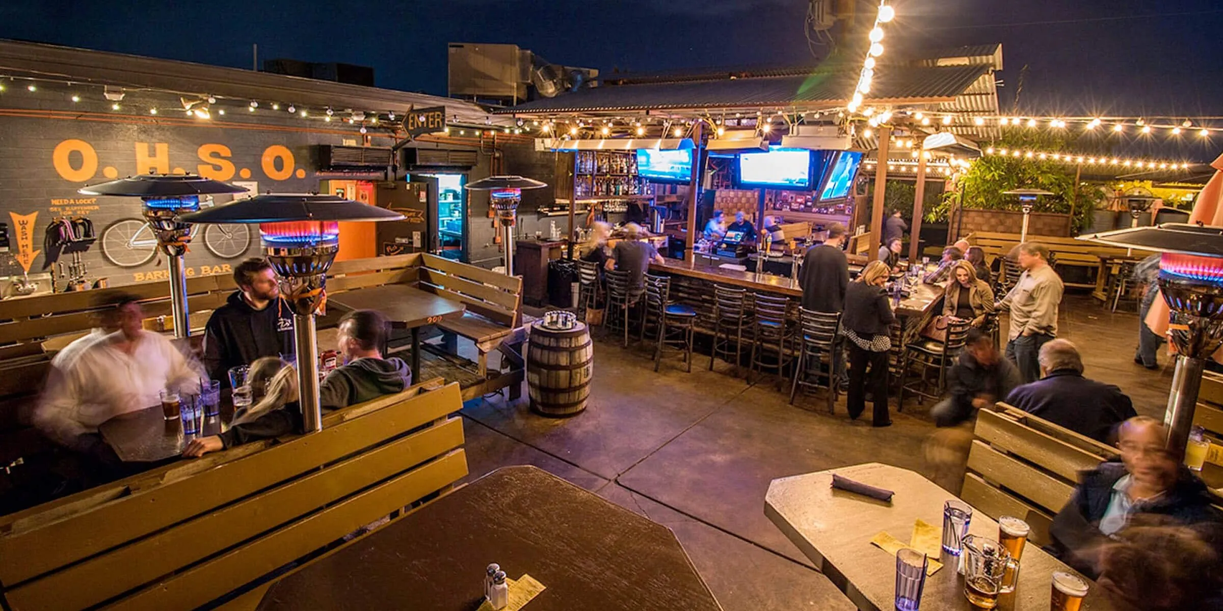 Outdoor brewery patio with heaters, string lights, wooden tables, and people socializing at night.