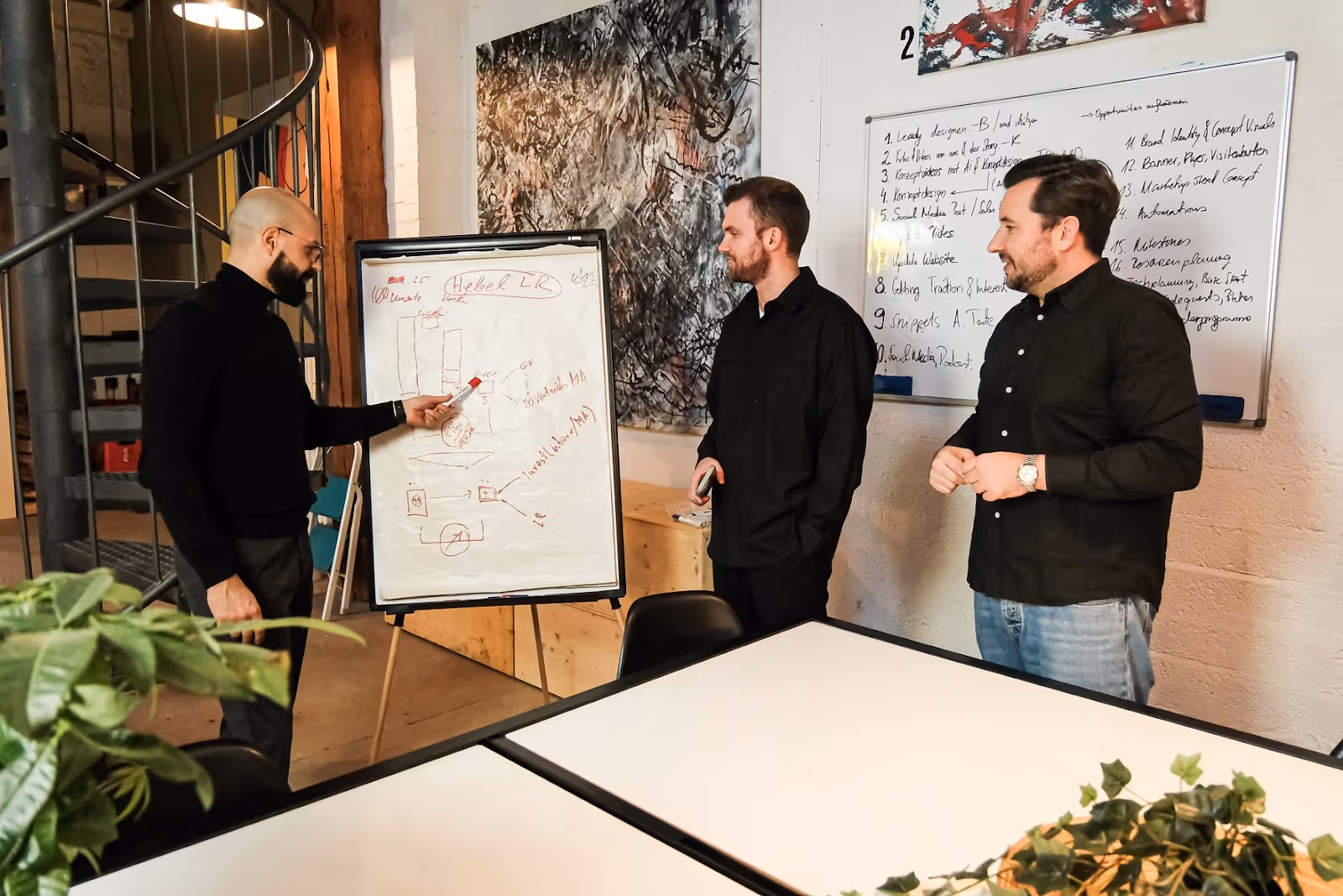 Three men in a casual office setting having a discussion around a presentation board with charts and notes.