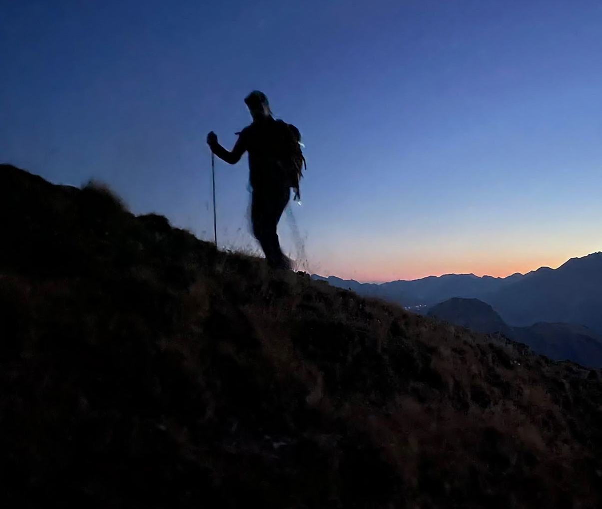 Silhouette d'un randonneur avec un sac à dos et un bâton marchant sur une pente en montagne au crépuscule.