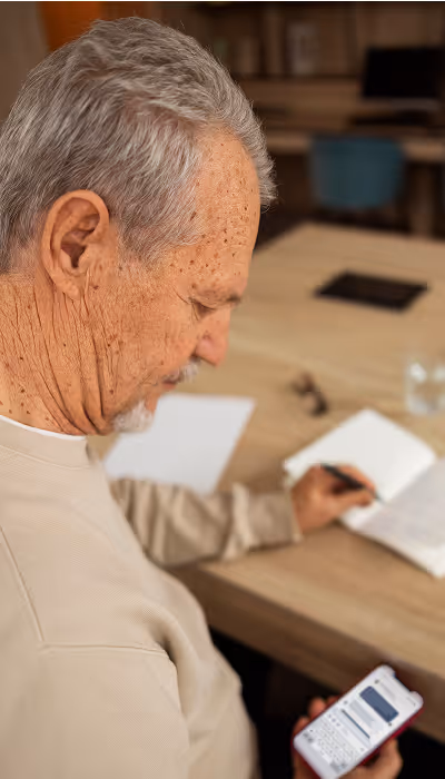 Älterer Mann mit grauen Haaren liest Textnachrichten auf einem Smartphone an einem Schreibtisch mit offenem Notizbuch.