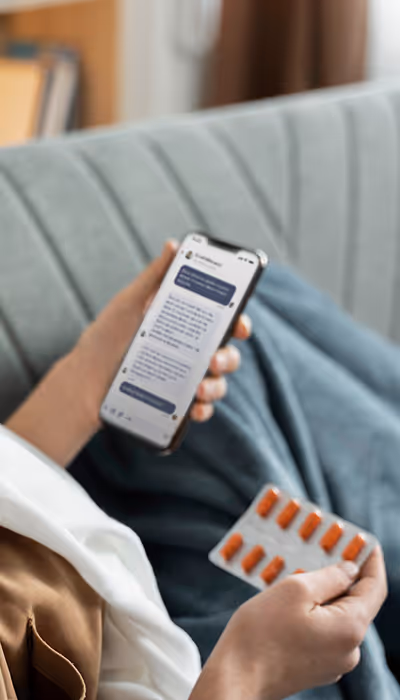 Osoba trzymająca smartfon wyświetlająca czat w aplikacji mobilnej Doctor.One i blister pomarańczowych kapsułek podczas siedzenia na kanapie.