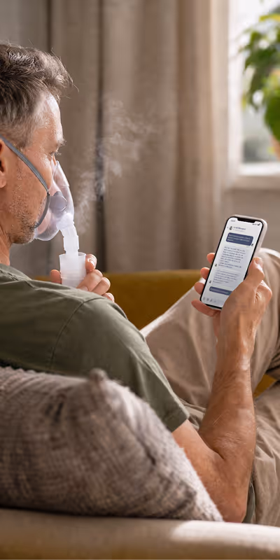 Man sitting on couch using a nebulizer mask while reading messages on his smartphone.