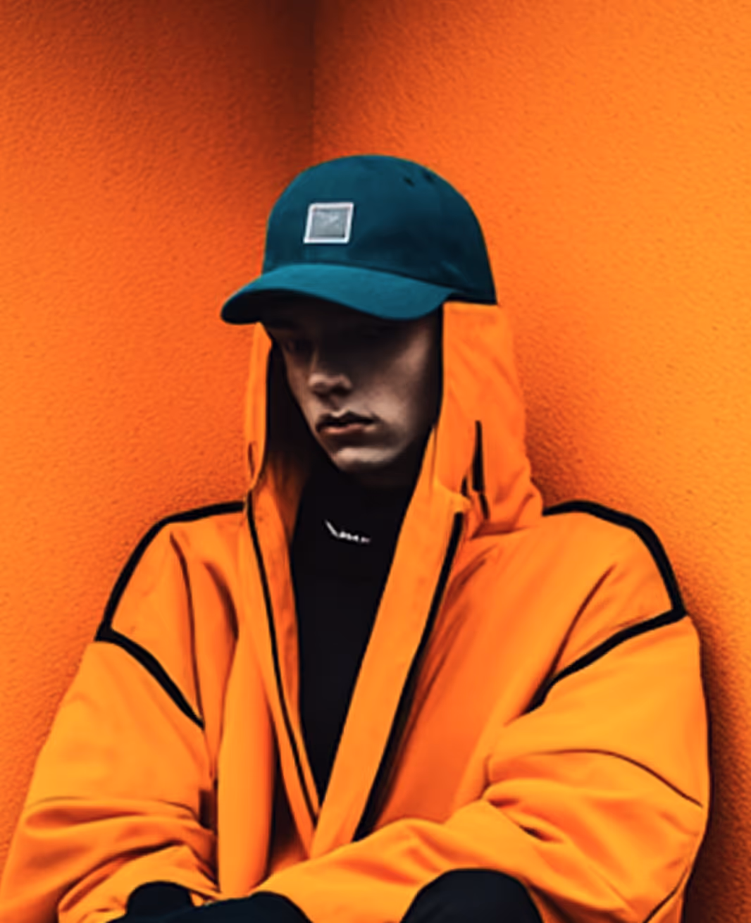 Person wearing an orange jacket with black accents and a teal cap, sitting against an orange wall looking down.
