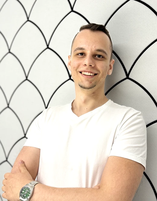 Smiling man with short hair wearing a white t-shirt and a silver watch, standing against a white wall with black scallop pattern.