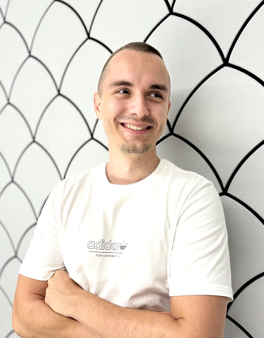 Smiling man with short hair wearing a white Adidas t-shirt standing with arms crossed against a white wall with black curved lines.