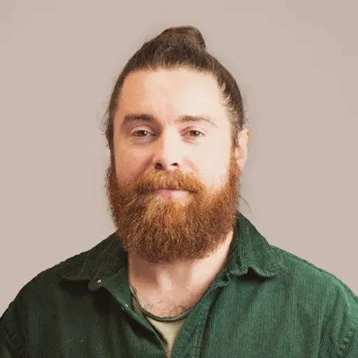 Man with a red beard and hair in a top bun wearing a green shirt, against a plain beige background.