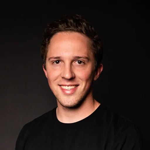 Young man with short brown hair smiling against a black background, wearing a black shirt.