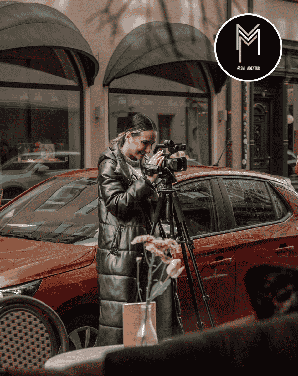 Woman in oversized white knitted sweater and pants crouching on snow-covered rock with snowflakes in her hair and wearing black sunglasses against a snowy mountain background.