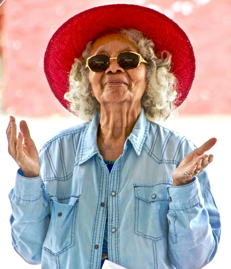 Stylish elderly person in red hat, sunglasses, and denim shirt gesturing