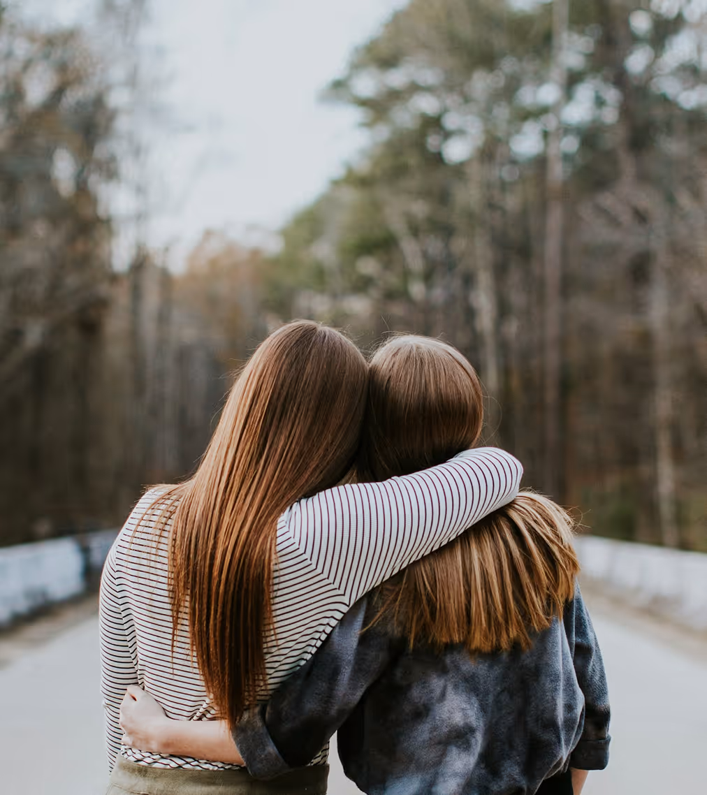 Due ragazze con i capelli lunghi si abbracciano camminando all'aperto su una strada con alberi sullo sfondo.