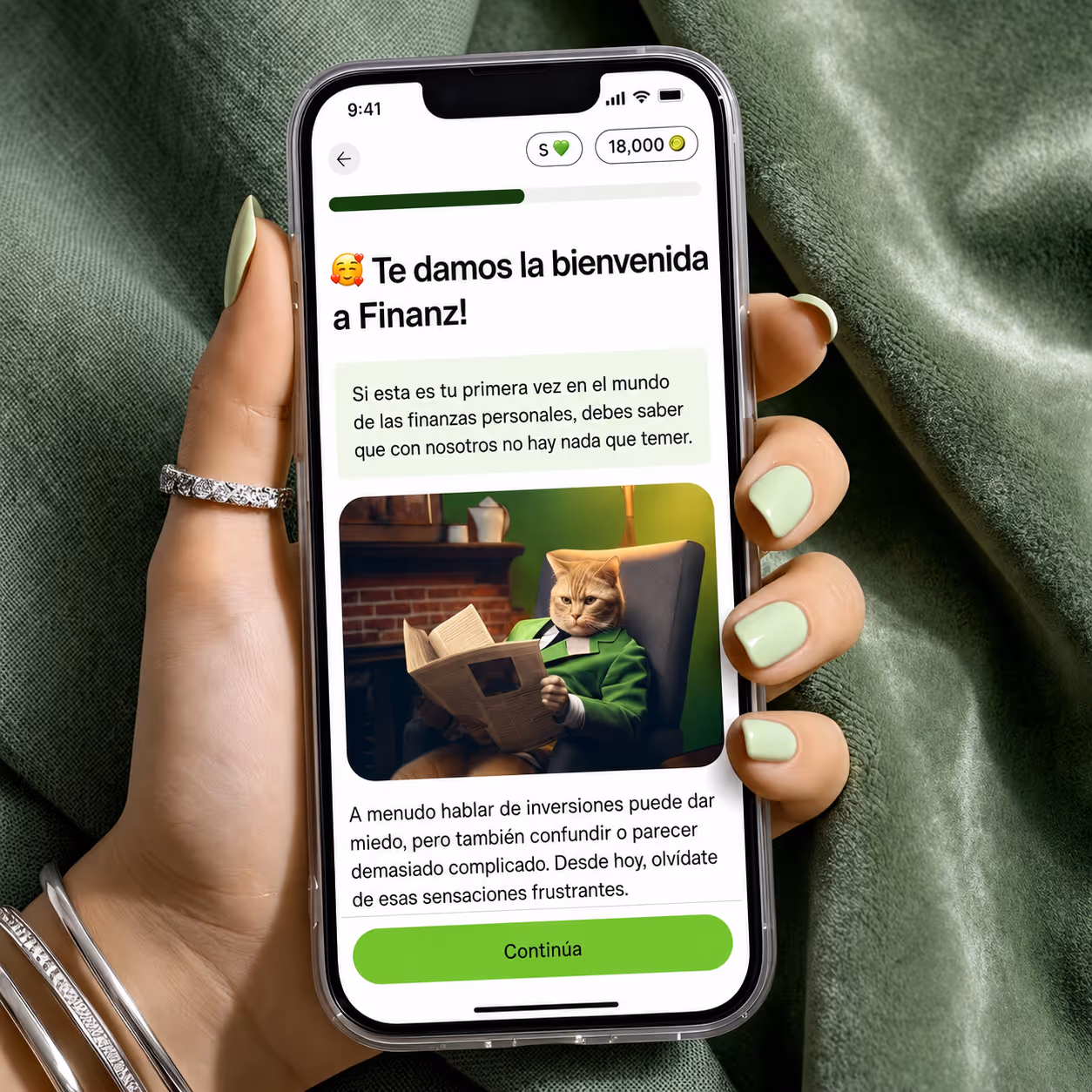 Una mano con esmalte de uñas verde claro sostiene un teléfono inteligente que muestra una aplicación de finanzas personales en italiano con la imagen de un gato disfrazado de hombre leyendo un periódico.