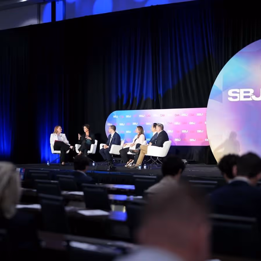 Two men seated on stage in discussion, surrounded by pink and blue backgrounds with text "SBJ CAA". One holds a microphone.