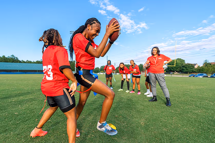 A group of women playing flag football. One holds a football, smiling, while others cheer. They're on a grassy field wearing red jerseys.