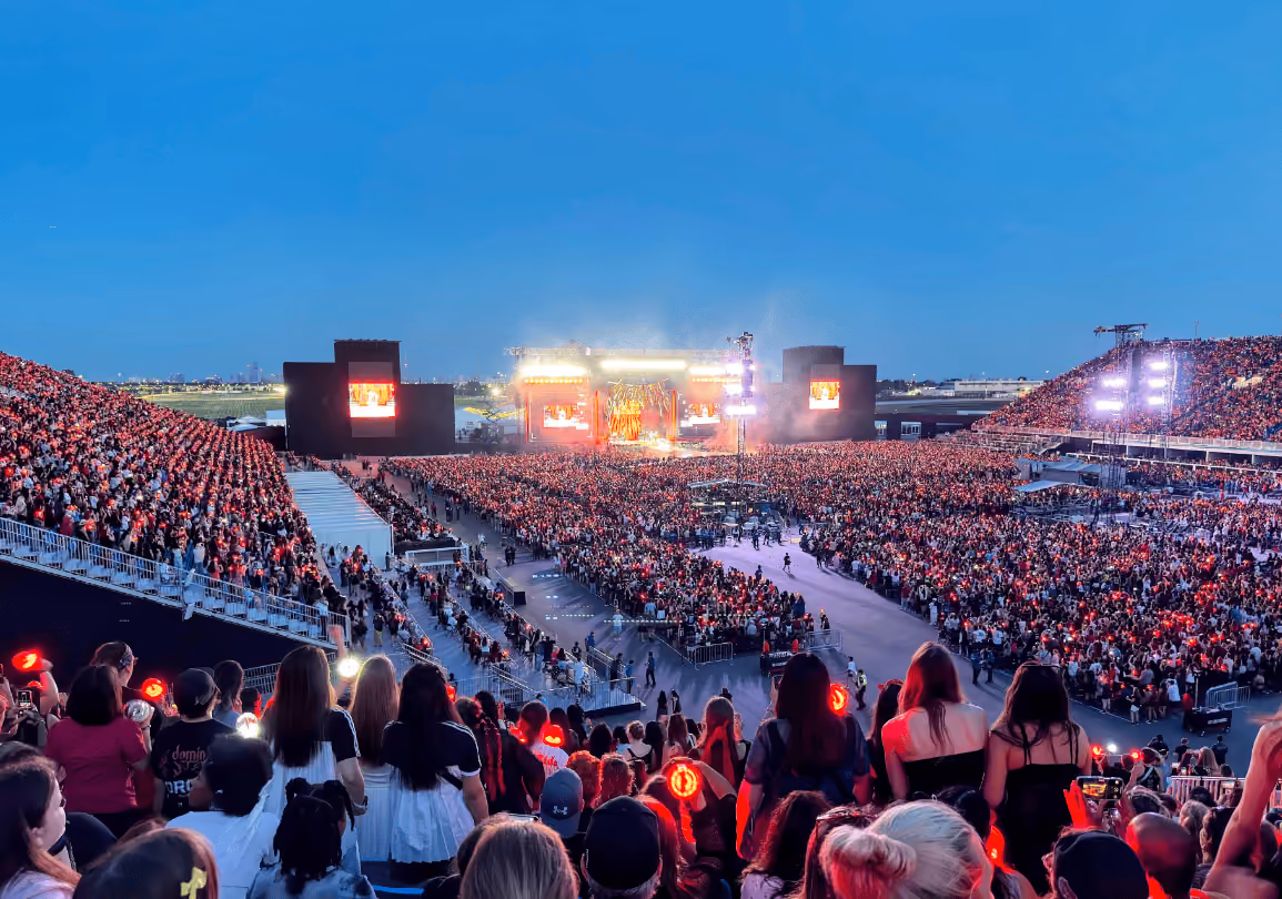 Massive outdoor concert crowd in a temporary stadium at dusk, facing a brightly lit stage under a blue sky, with red glow sticks.