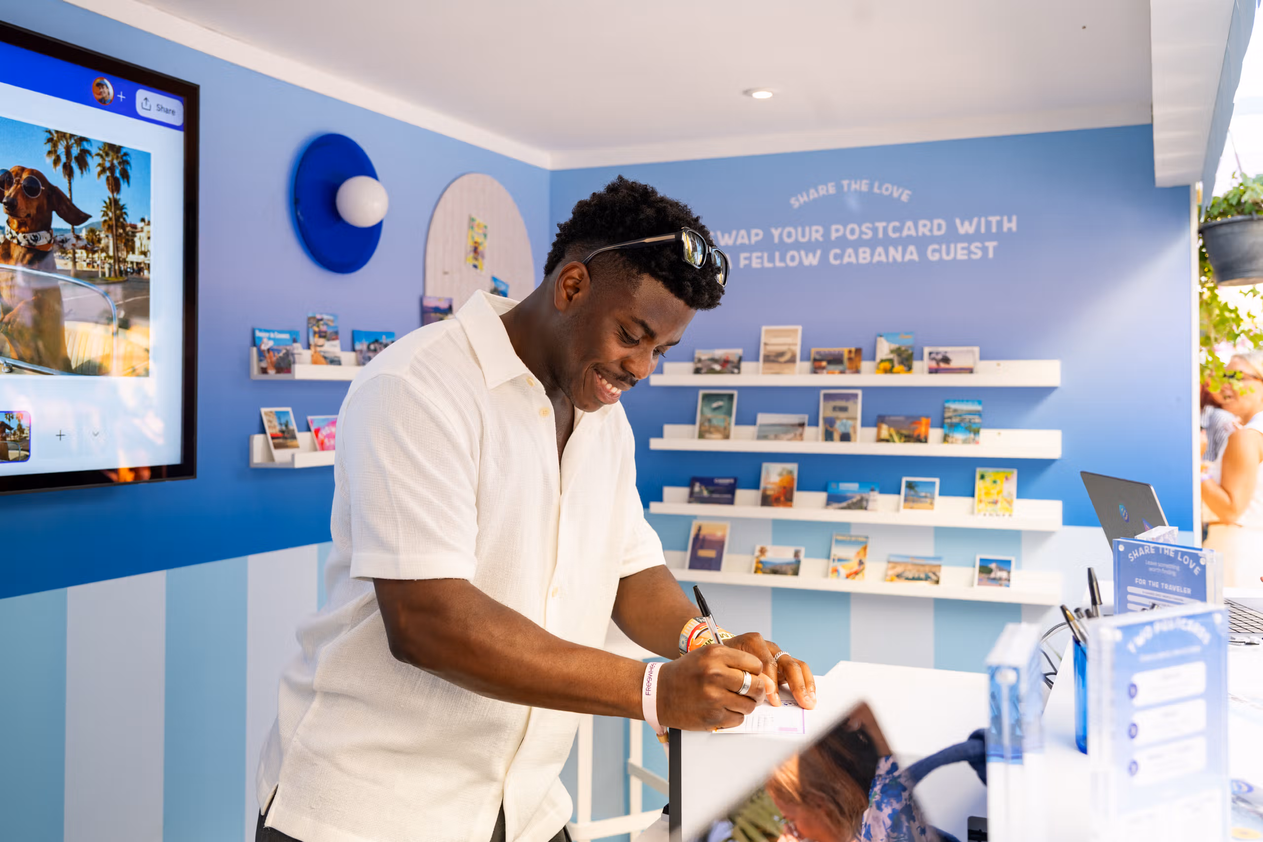 A guest writing a postcard at the Canva Postcard Parlour, with racks of printed postcard designs on the blue wall behind him.