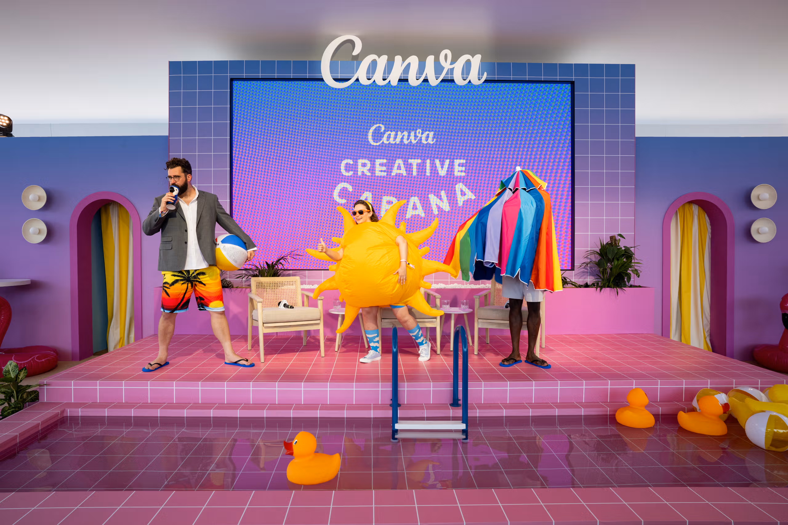 Guests on the Canva Creative Cabana pool stage at Cannes, one wearing an inflatable sun costume, with rubber ducks floating in the pink tiled pool below.