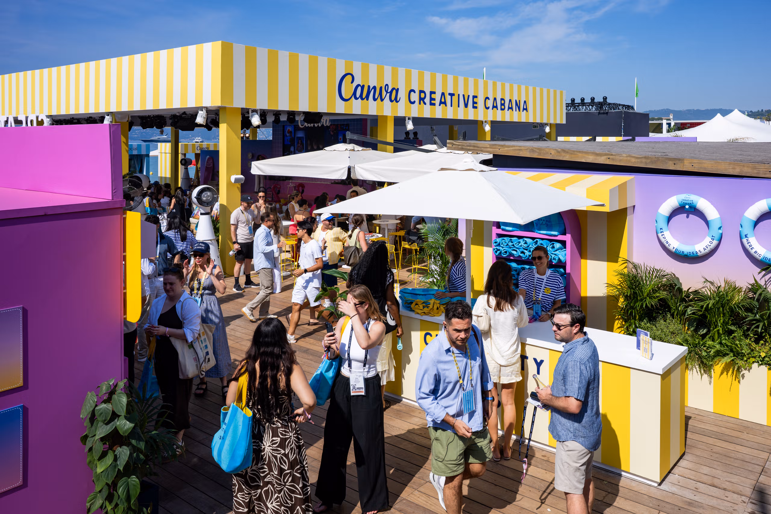 Guests walking the boardwalk beneath the yellow striped Canva Creative Cabana awning at Cannes, past white parasols and pink and purple structures.