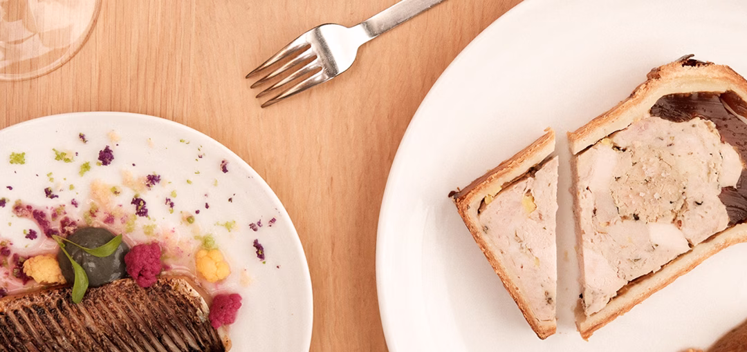 Deux assiettes sur une table en bois, l'une avec une tranche de pâté en croûte coupée en deux, l'autre avec un morceau de viande grillée accompagnée de petits légumes colorés.