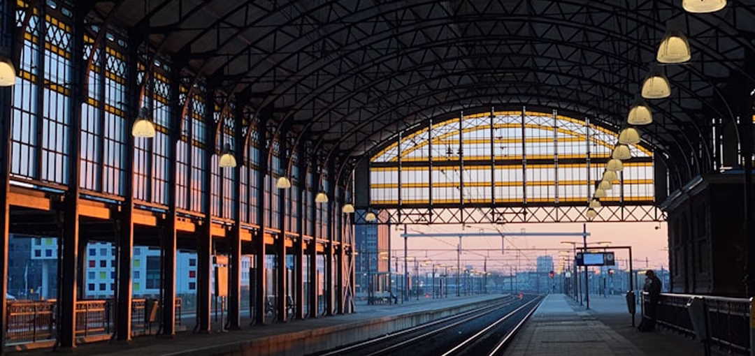 Intérieur d'une gare avec une grande verrière en fer et des rails de train s'étendant au loin au coucher du soleil.