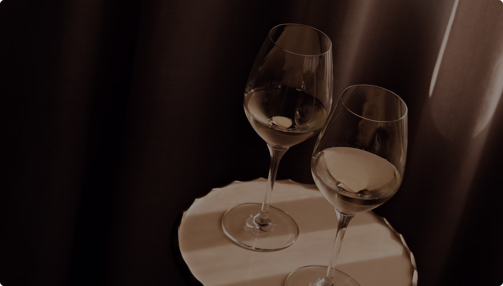 Deux verres à vin blanc posés sur une table ronde éclairée, avec un fond de rideaux sombres.