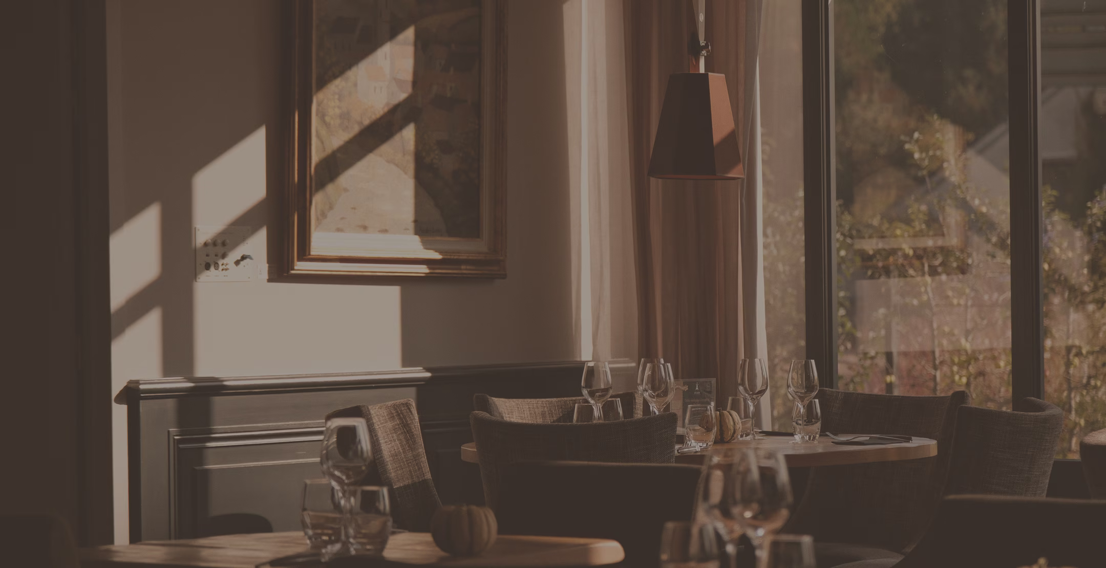 Intérieur de restaurant avec tables dressées de verres, chaises confortables, et une grande fenêtre laissant entrer la lumière naturelle.