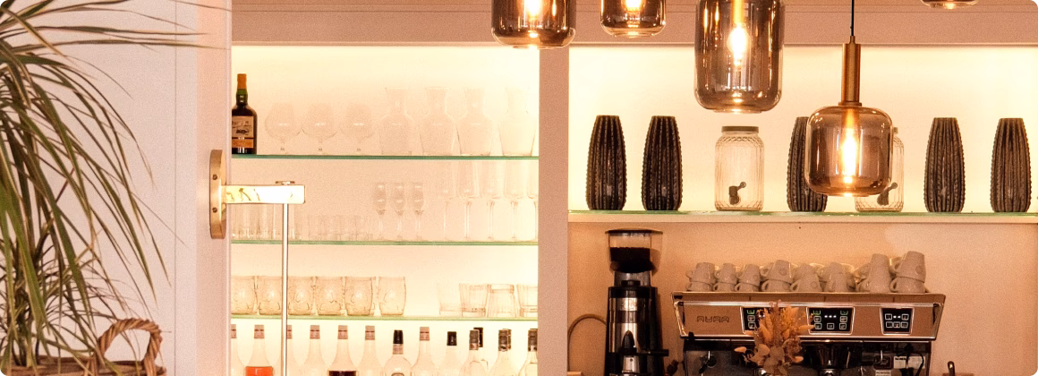 Intérieur de bar avec estantes lumineuses affichant des verres, bouteilles, et une machine à café sous des suspensions en verre teinté.