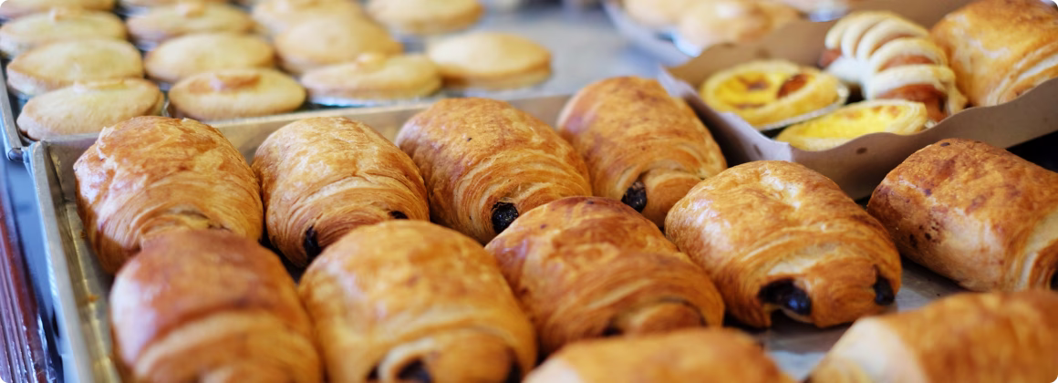 Une variété de viennoiseries françaises fraîches, comprenant des pains au chocolat et des tartes aux œufs, disposées sur des plateaux.