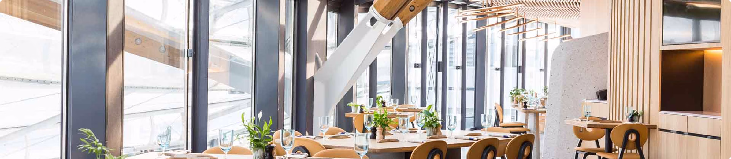 Intérieur de restaurant moderne avec grandes fenêtres, tables en bois, chaises jaunes et plantes décoratives.