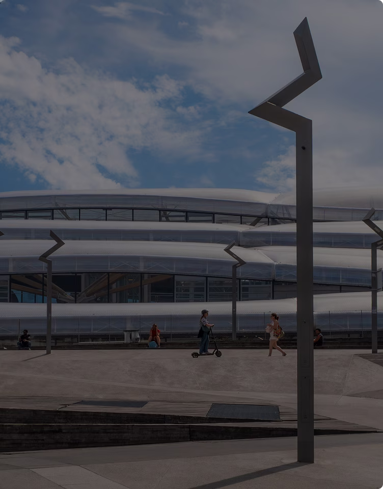 Un homme sur une trottinette et une femme marchant devant un bâtiment moderne avec une façade en métal ondulé, sous un ciel partiellement nuageux.