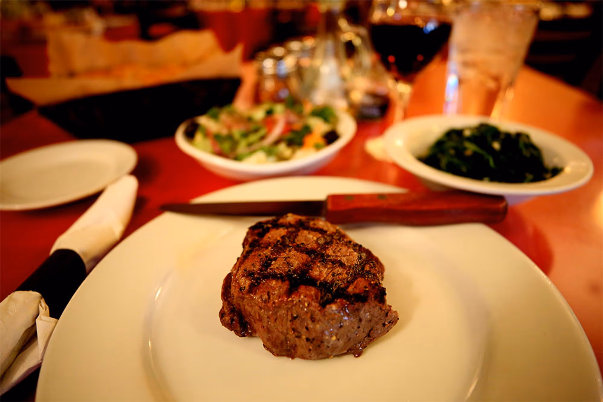Grilled steak on a white plate with a steak knife, bowl of salad, sautéed greens, glass of red wine, and glass of water on a restaurant table.