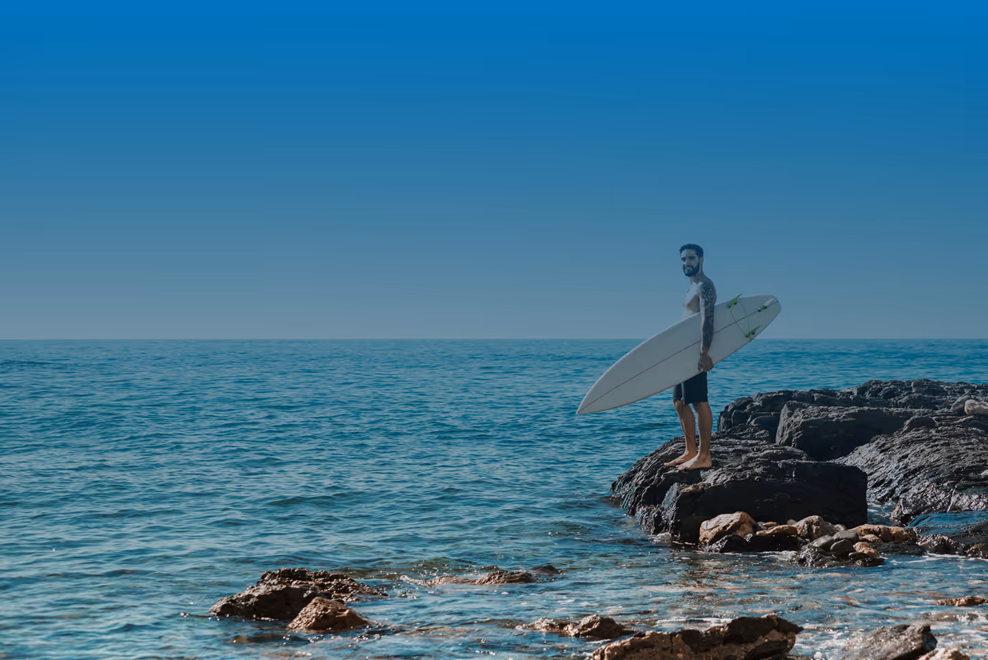 Man standing barefoot on rocky shore holding a white surfboard looking at the ocean.