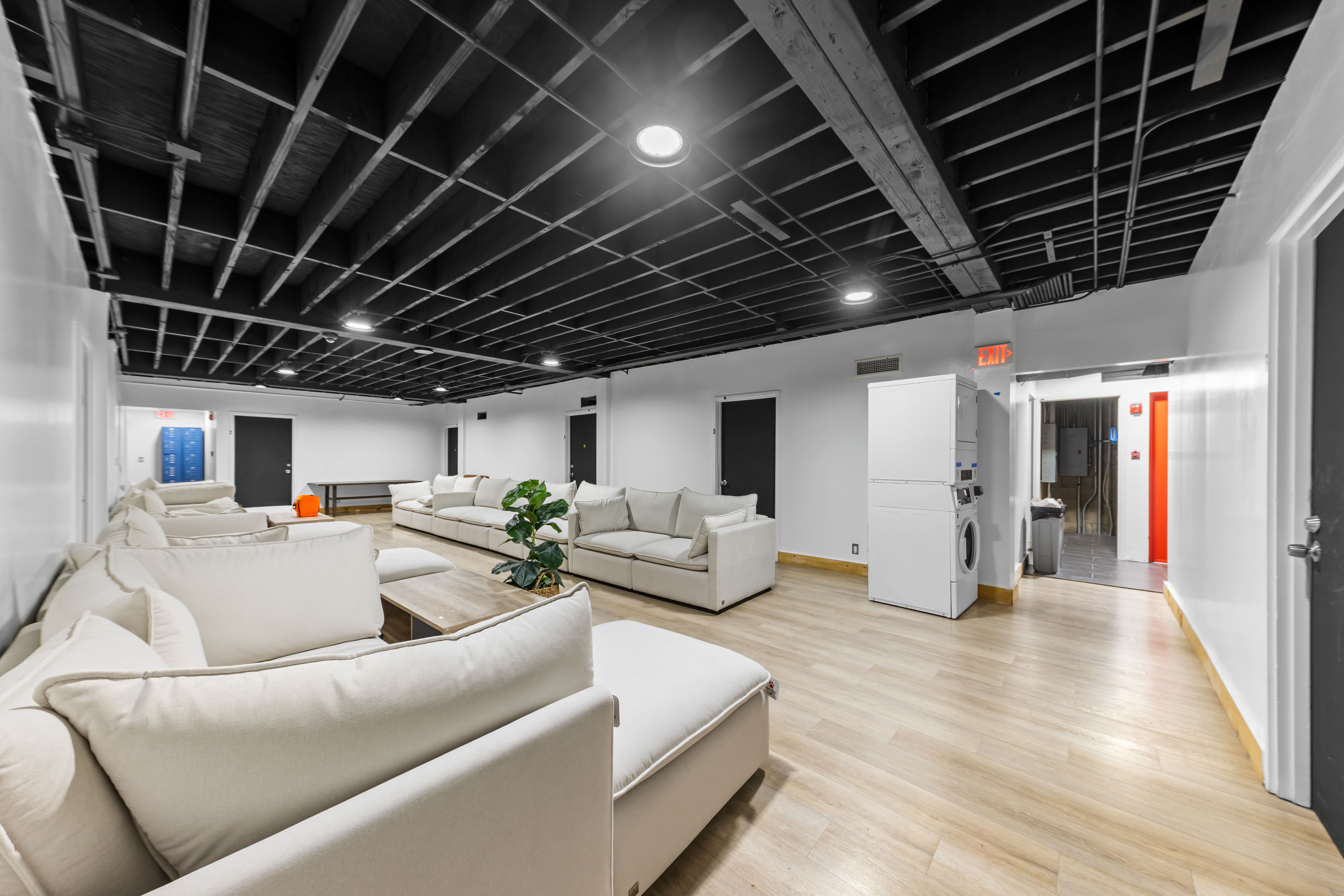Room with beige sofas, wooden floors, black ceiling beams, a plant, and stacked washer and dryer.