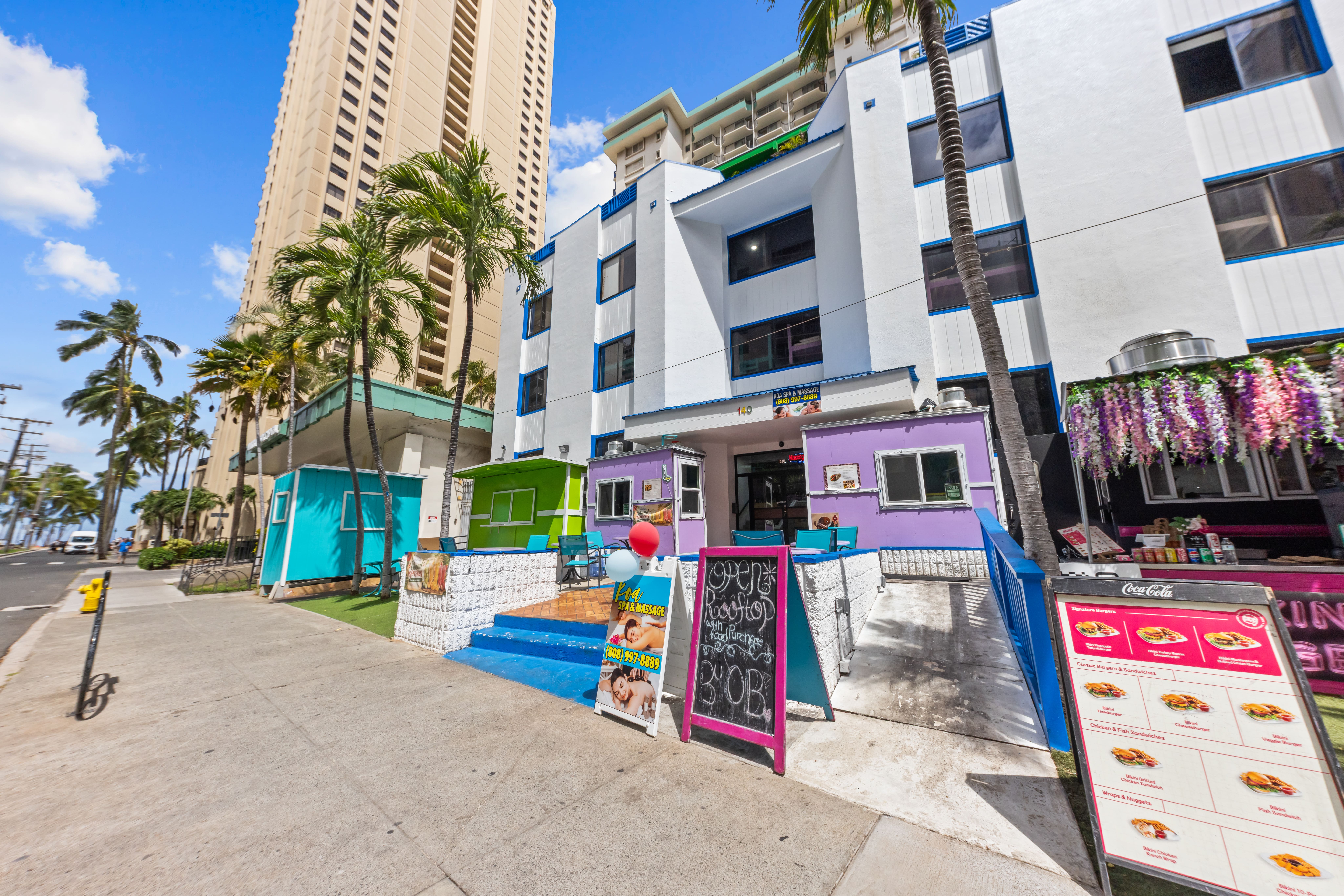Aerial view of a sandy beach with palm trees and a walking path next to clear ocean water, with high-rise buildings and cloudy sky in the background.