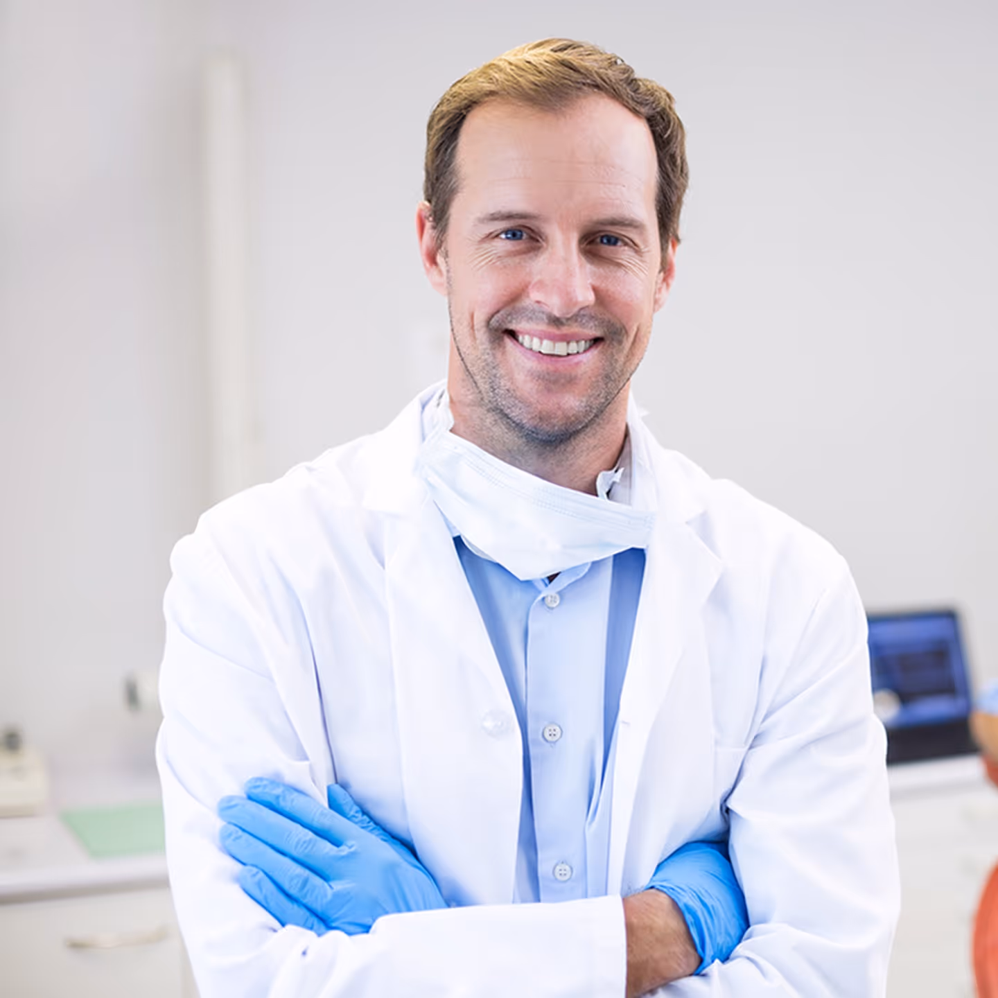 Smiling male dentist in a white coat and blue gloves with arms crossed in a medical office.