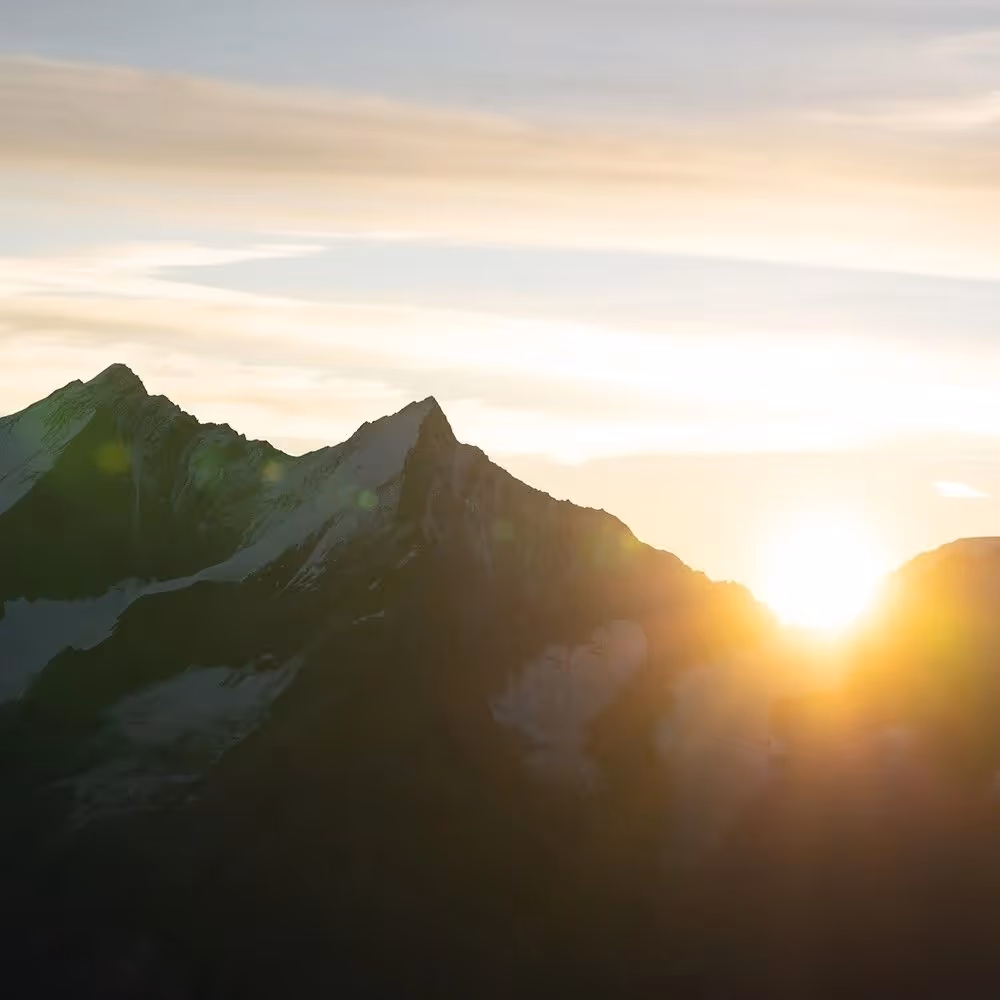 Sun rising behind snow-capped mountain peaks under a partly cloudy sky.