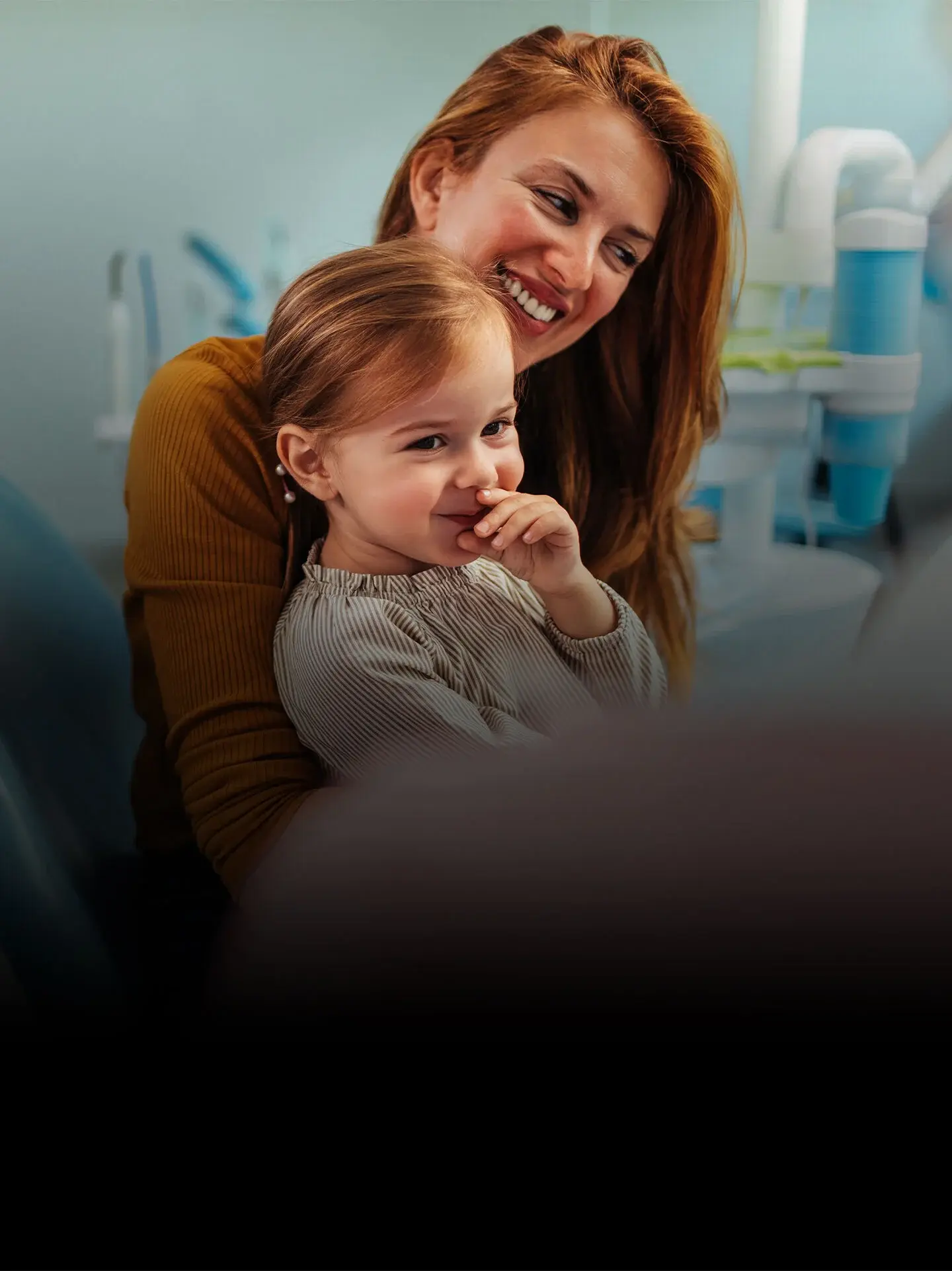 Smiling woman with red hair hugs a young girl who is covering her mouth and looking to the side, in a medical or dental office setting.