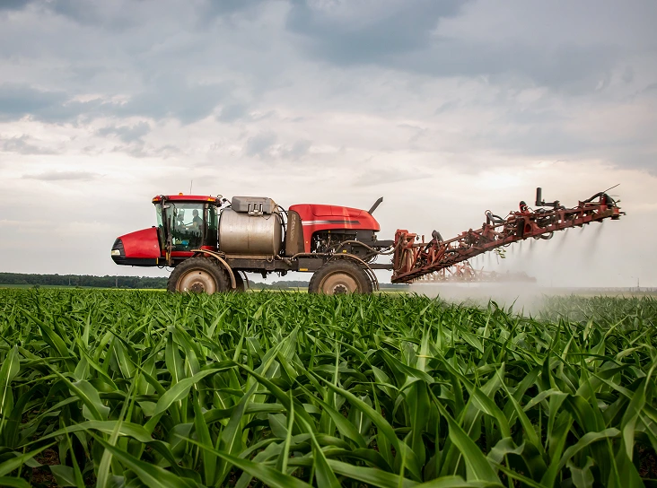 Red agricultural sprayer vehicle spraying crops in a vast green field under a cloudy sky.