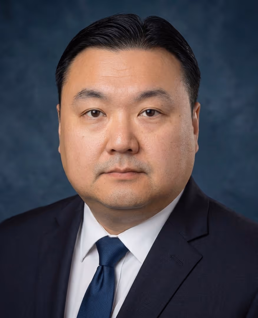Serious man in dark suit, white shirt, and blue tie with dark hair combed back against blue background.